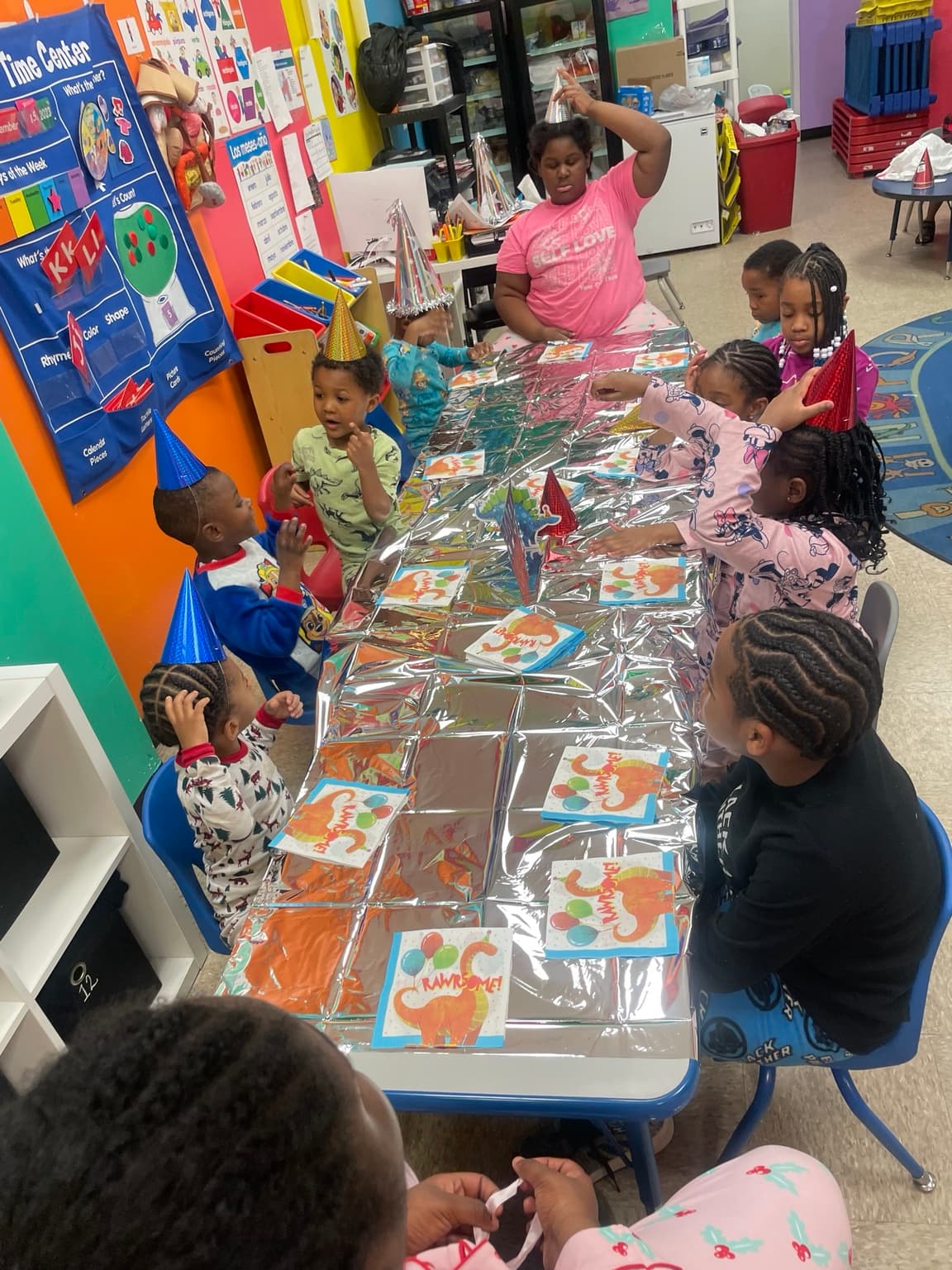 A teacher wearing a party hat sits with children in pajamas at a decorated table during a classroom celebration
