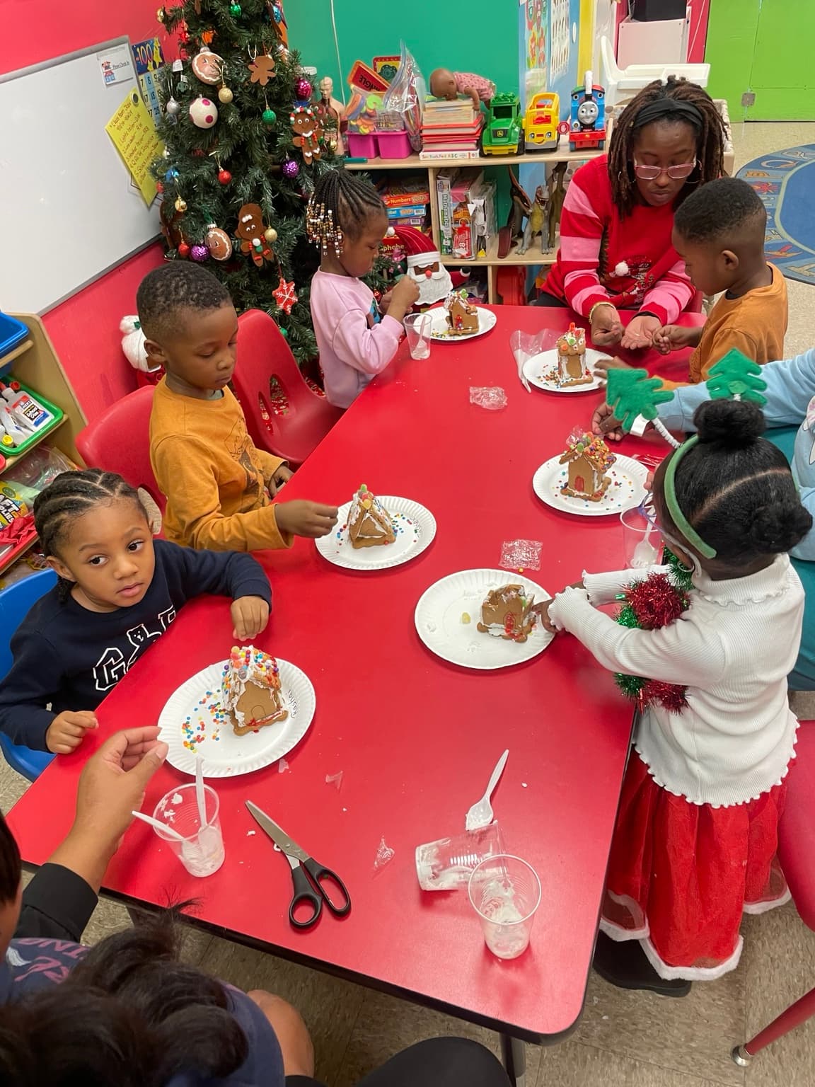 A teacher helps children decorate gingerbread houses at a table beside the classroom Christmas tree