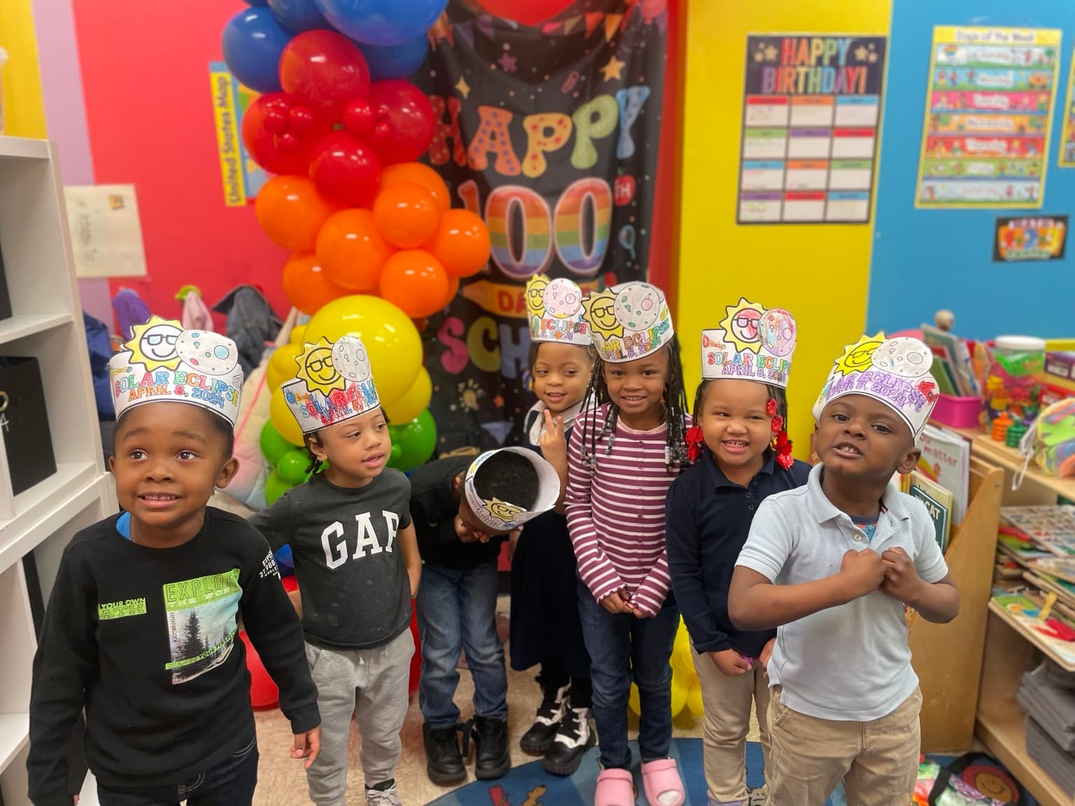 A group of children wearing handmade solar eclipse hats posing together in front of a 100 Days of School balloon display
