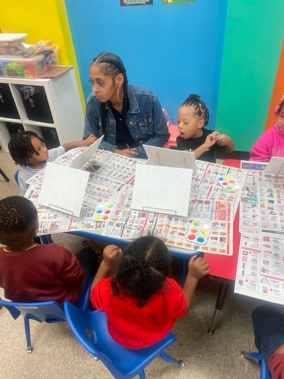 A teacher helps children paint at easels reading 'I Love Art' during a classroom art lesson