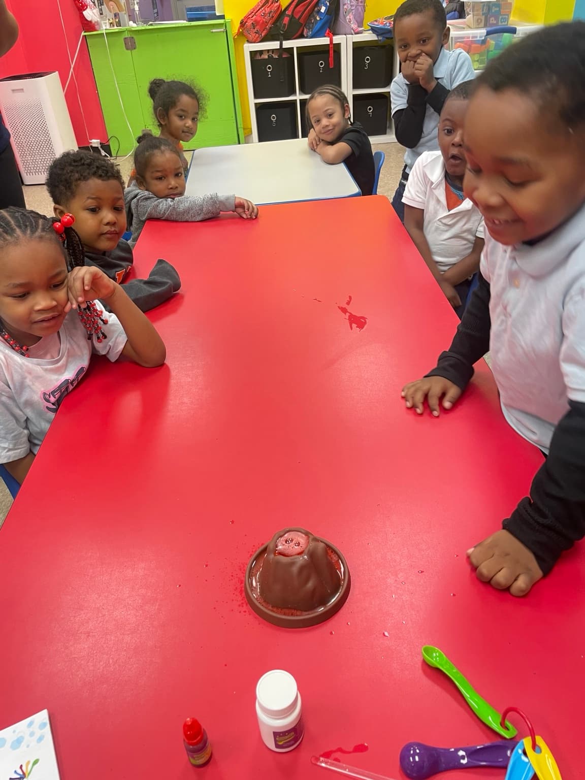 Children gathered around the table in wonder watching a baking-soda volcano experiment erupt
